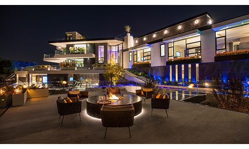 A luxurious modern house with extensive outdoor landscape lighting at night. The well-lit patio features a circular fire pit surrounded by seating, with the house showcasing multiple levels and large windows.
