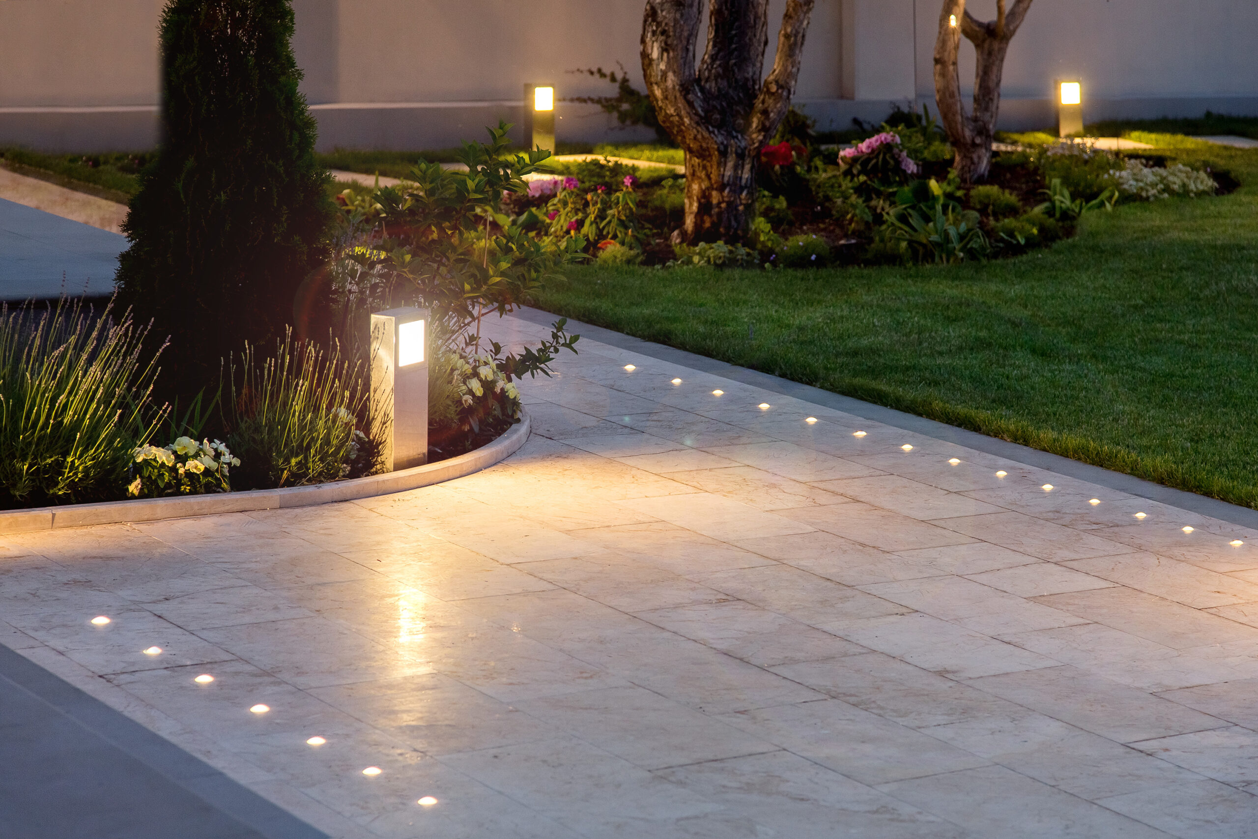 A modern outdoor garden pathway featuring sleek tile flooring with embedded LED lights for illumination. The path is bordered by well-maintained greenery and subtle automated bollard lights, creating an elegant and functional lighting solution. The surrounding garden includes vibrant flowers and neatly trimmed grass, enhancing the inviting atmosphere of the space.