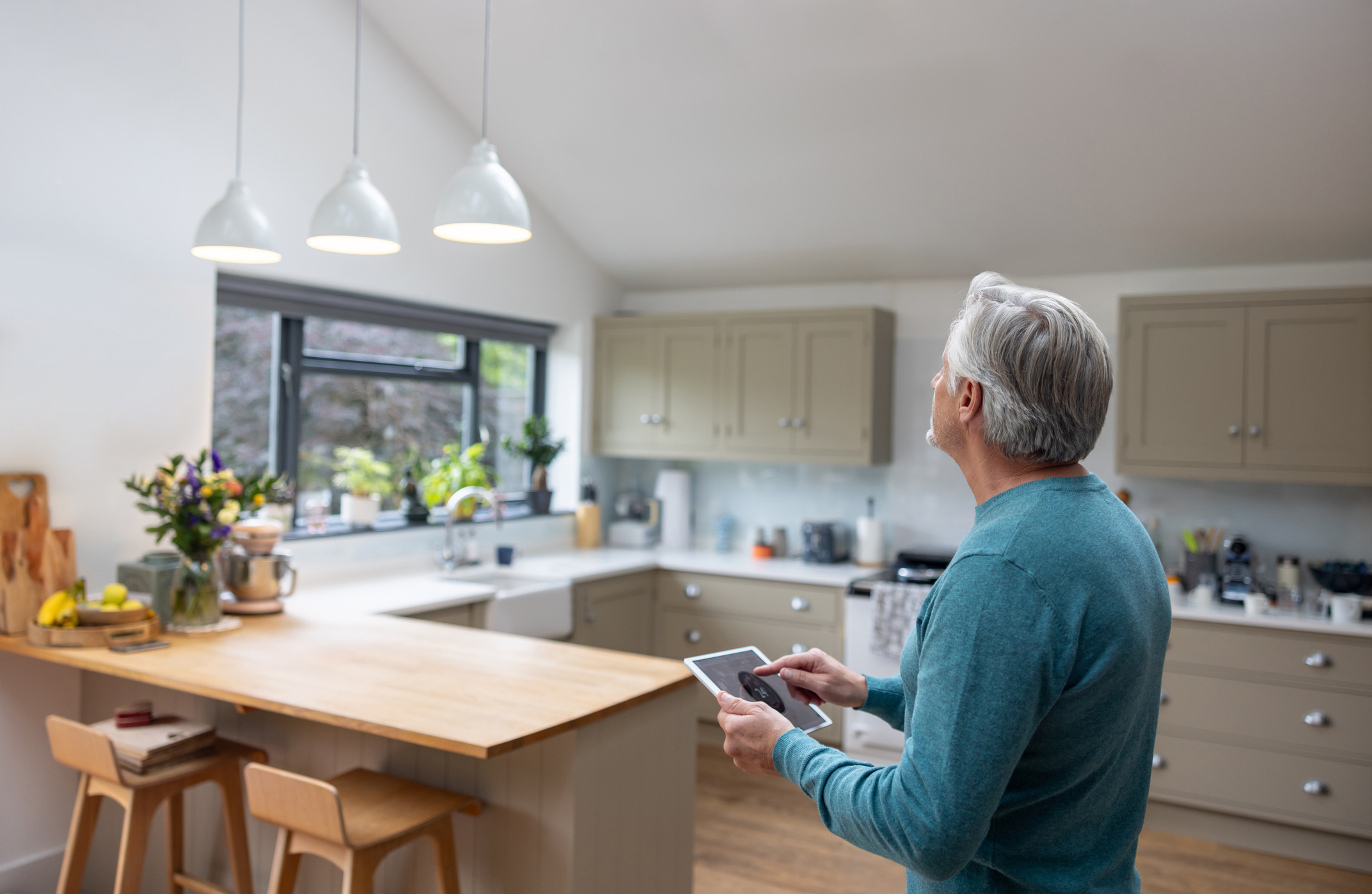 A middle-aged man standing in a modern, brightly lit kitchen, holding a tablet and interacting with a home automation system, possibly controlling the three pendant lights hanging above a wooden kitchen island. The kitchen is furnished with beige cabinets, a sink with a view to an outside garden, and various kitchen items neatly arranged, reflecting a well-maintained, contemporary, and automated home environment.