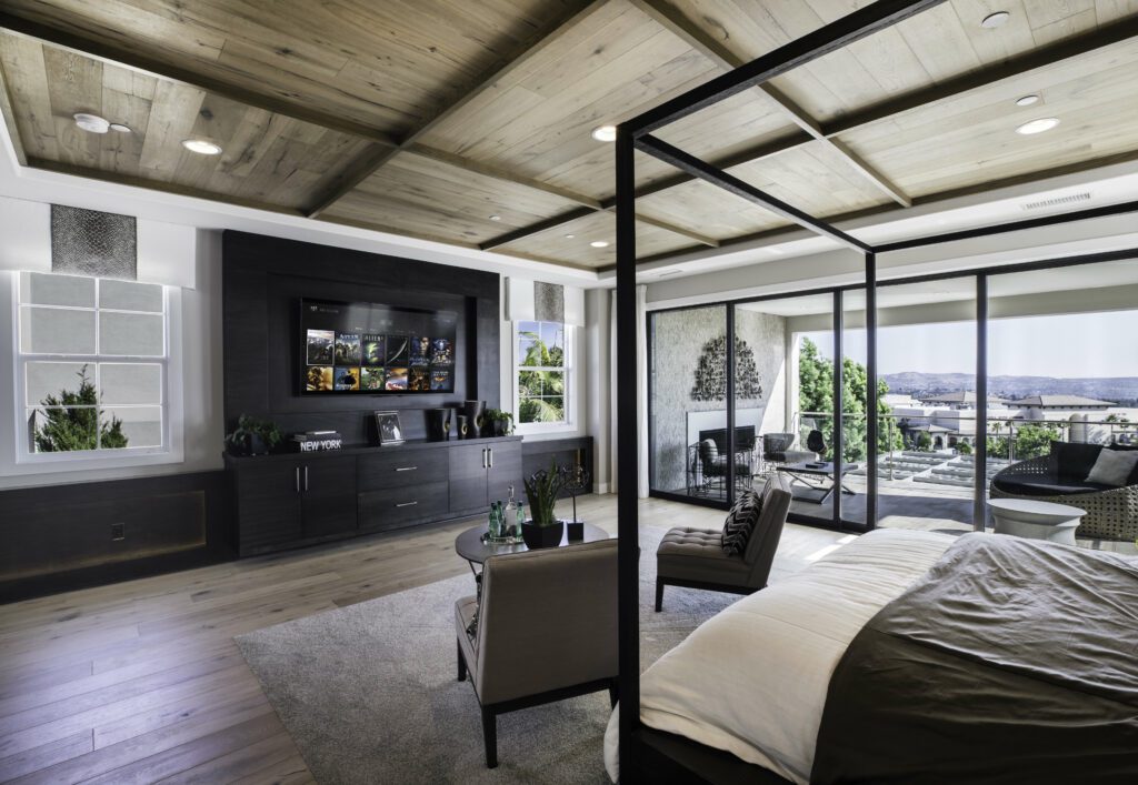 A luxurious modern bedroom with a four-poster bed, wooden ceiling, and floor-to-ceiling glass doors leading to a spacious balcony with scenic views fitted with automated shades. The room features a built-in entertainment center with a large TV, cozy seating, and stylish decor, blending contemporary and natural design elements.