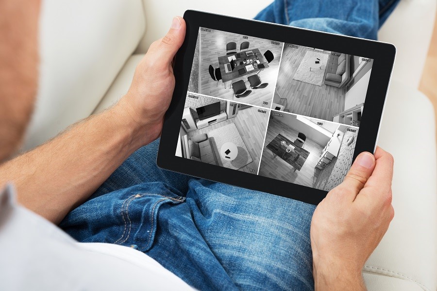 A person sitting on a couch, holding a tablet displaying a live feed of four black-and-white home security camera views. The tablet screen shows different rooms in a house, including a living room, dining area, and hallway, emphasizing home security monitoring and smart surveillance technology.