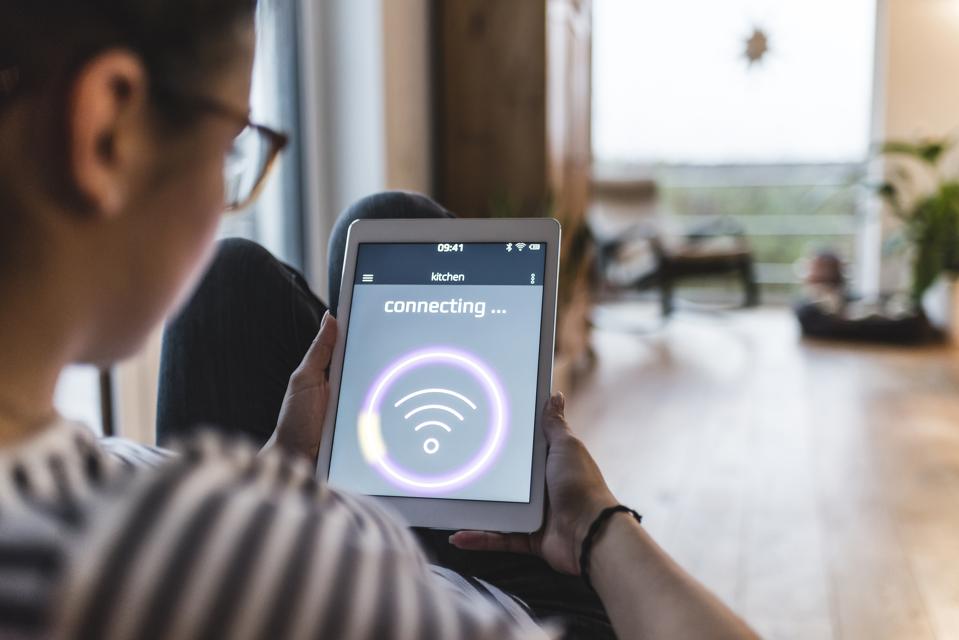 A person sitting in a modern home holding a tablet displaying a "connecting..." screen with a Wi-Fi symbol. The background features a well-lit, cozy living space with wooden floors and plants, emphasizing smart home connectivity and wireless internet access.