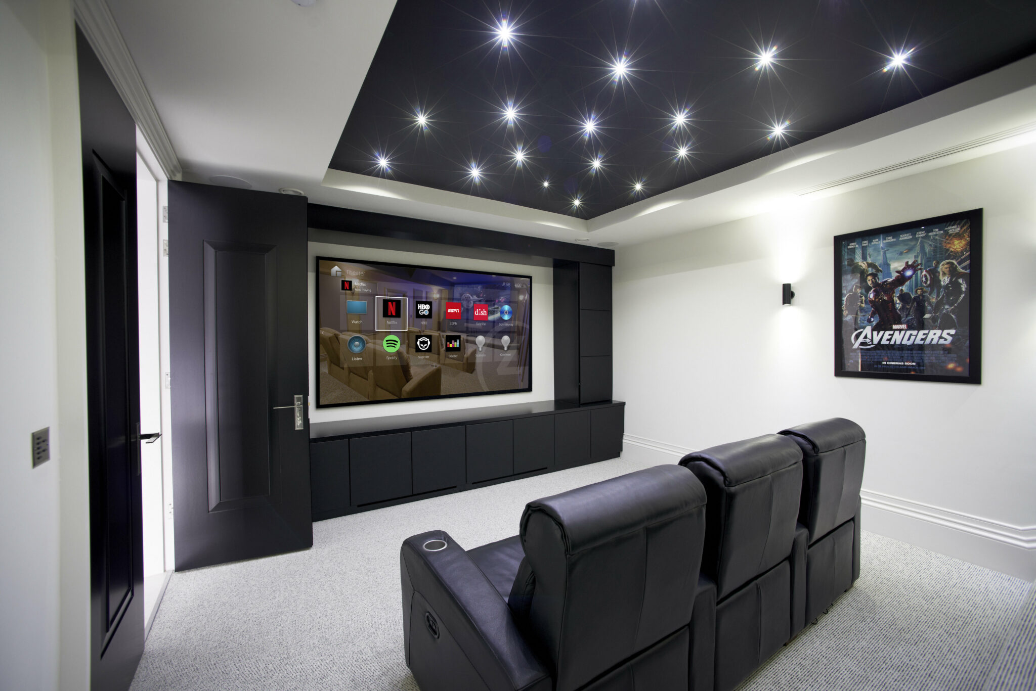 A sleek, modern home theater with black leather reclining seats facing a large wall-mounted screen displaying streaming apps and surround sound system. The ceiling features star-like recessed lighting, and a framed "Avengers" movie poster decorates the wall. The minimalist design and ambient lighting create an immersive entertainment experience.